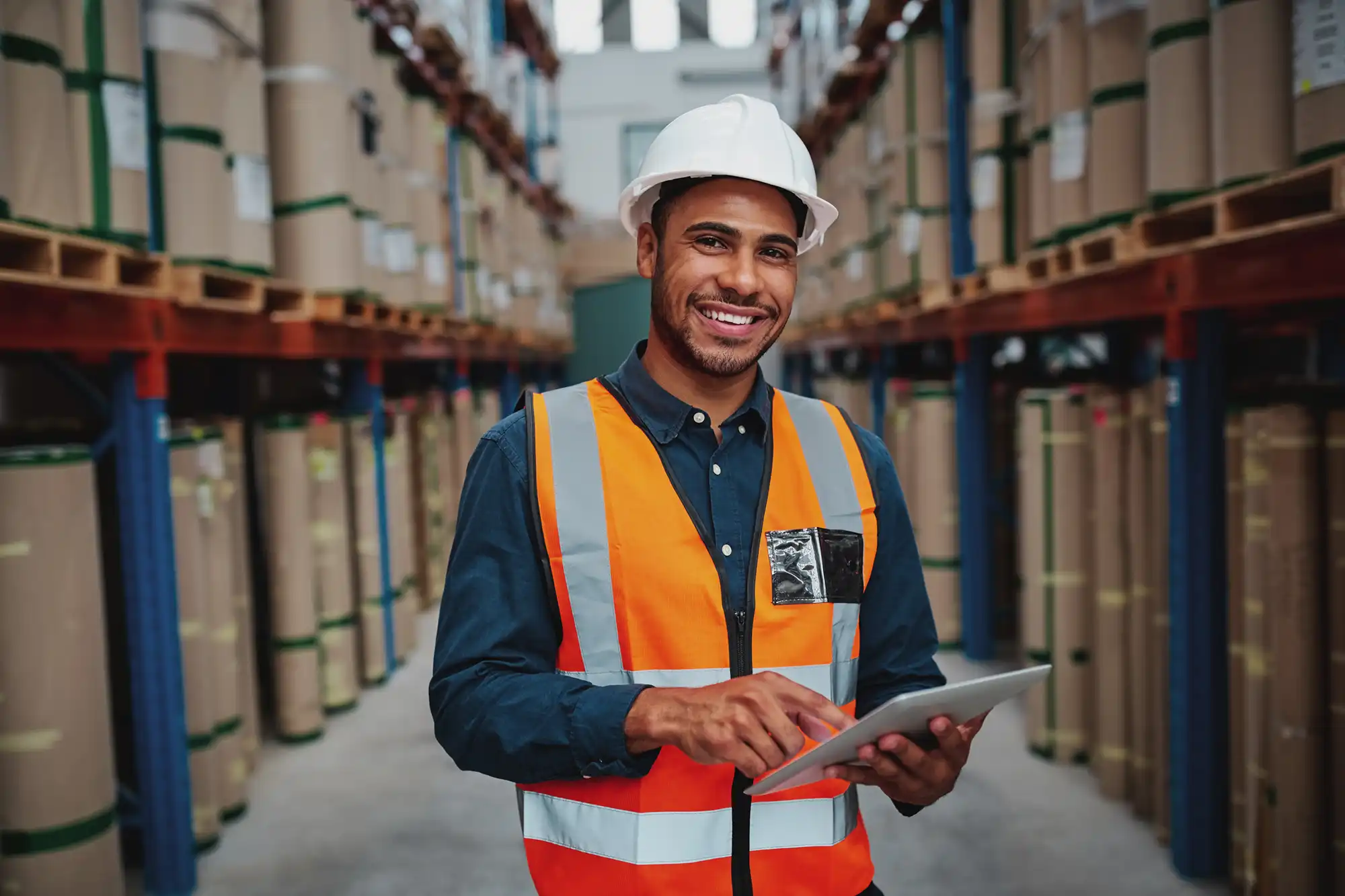 A man in a warehouse is smiling as he holds a tablet checklist. He is surrounded by boxes.